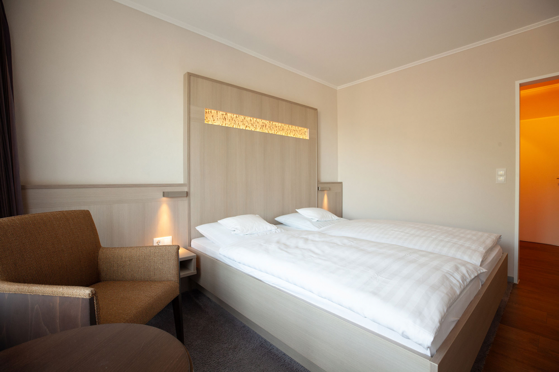 A modern hotel room with a large double bed, white bedding, an upholstered headboard, a brown armchair, a small round table and minimal décor. The warm lighting gives the neutral room a cosy atmosphere.
