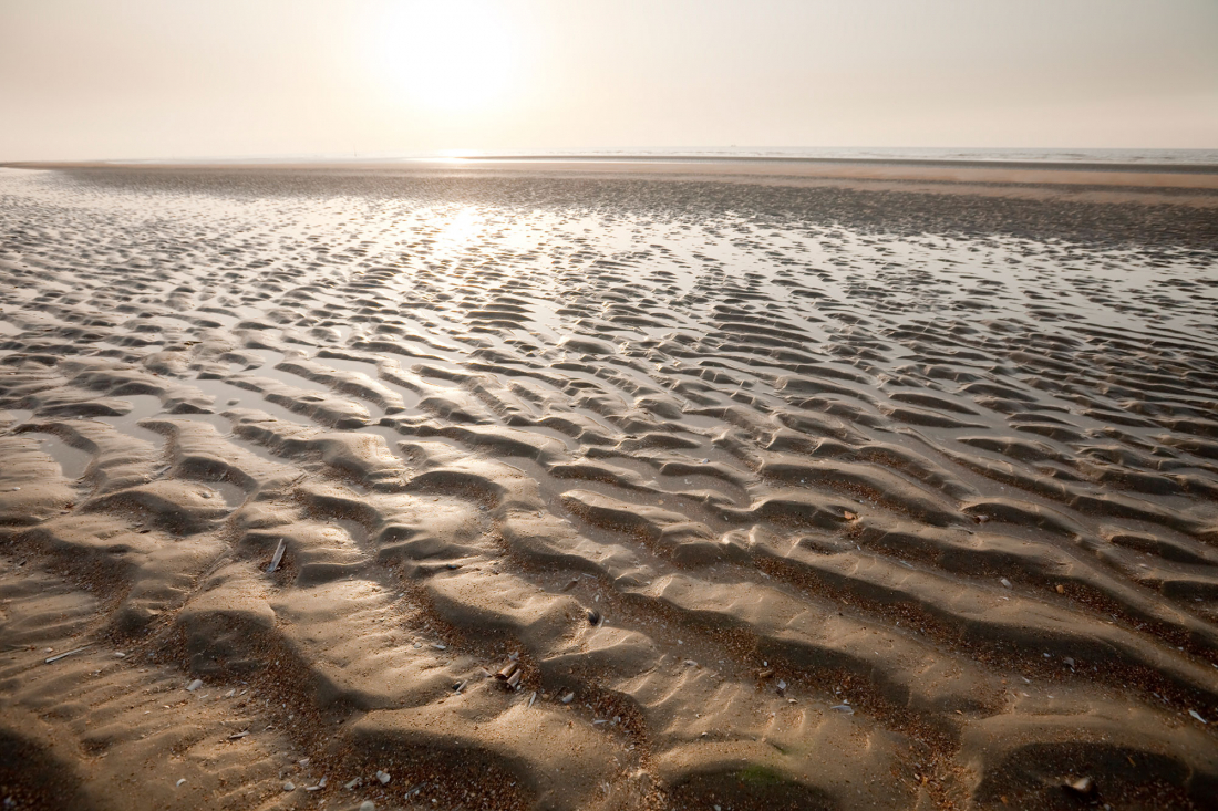Das Wattenmeer bei Ebbe mit seichten Wellen und Mustern im nassen Sand, in dem sich das helle Sonnenlicht in der Ferne unter einem dunstigen Himmel spiegelt.