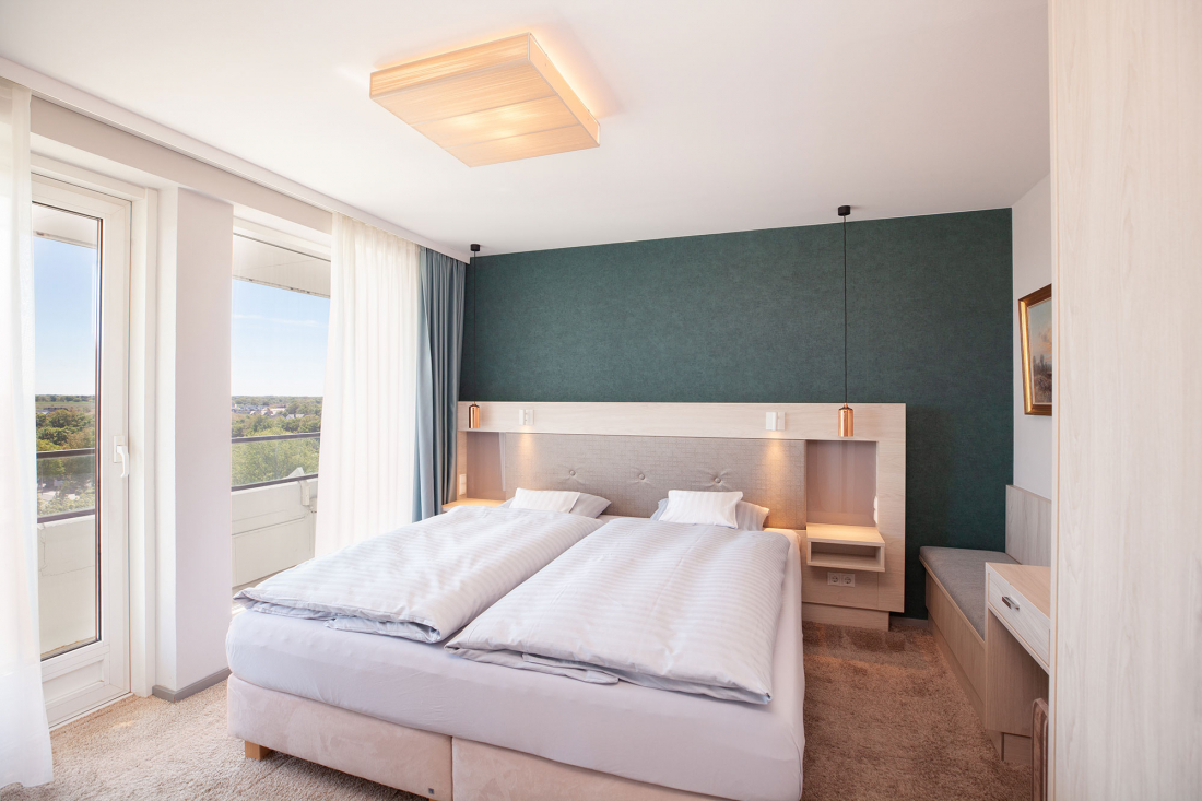 Ein modernes, helles Schlafzimmer mit einem Doppelbett, einer blaugrünen Akzentwand, einem eingebauten Kopfteil mit Regalen, sanfter Beleuchtung, einem großen Fenster mit transparenten Vorhängen und neutralen Möbeln.