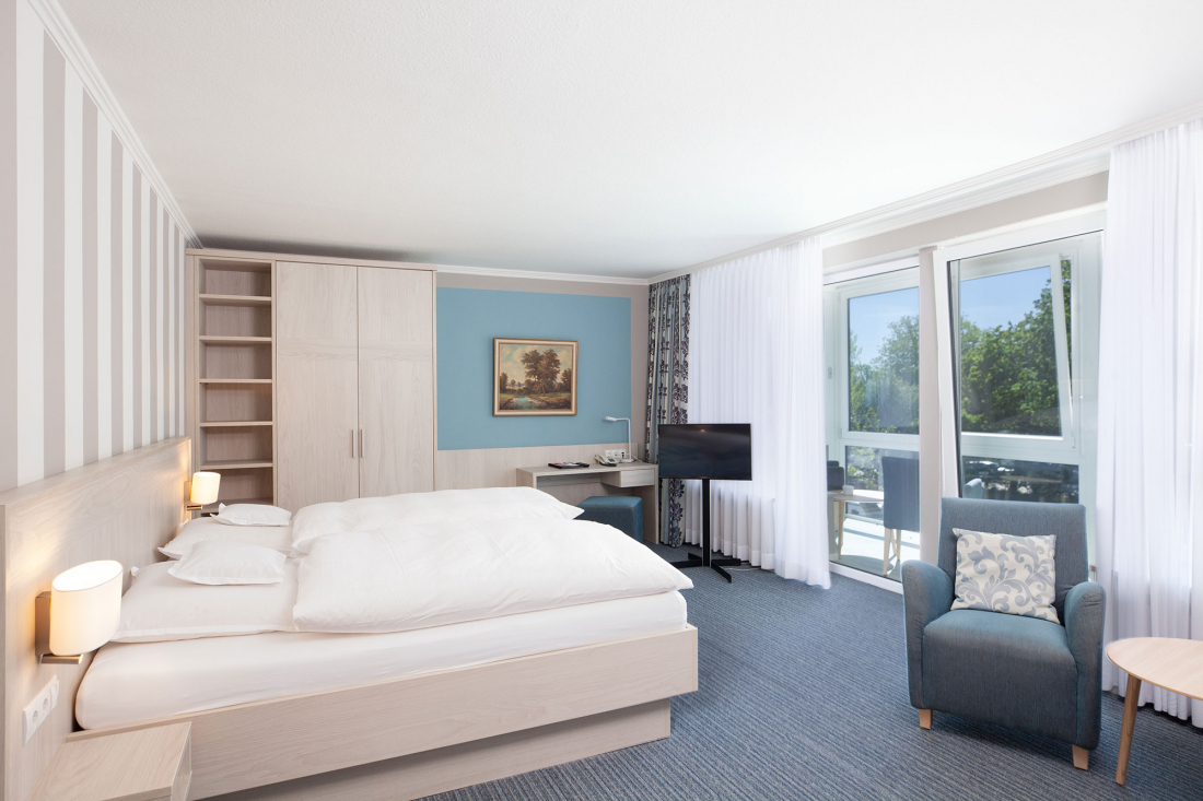 A bright hotel room with a double bed, armchair, desk, TV, large wardrobe and a window with sheer curtains overlooking the greenery. The furnishings are bright with blue and white colours and a painting on the wall.