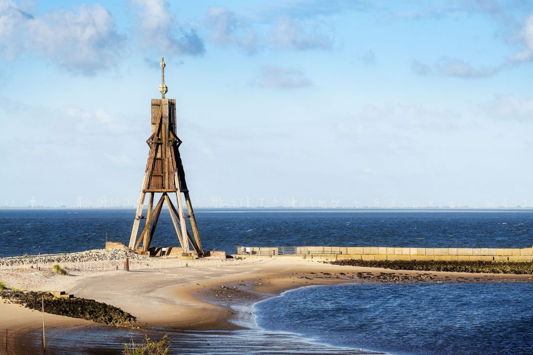 Das Wahrzeichen von Cuxhaven, die Kugelbake steht an einem sichelförmigen Sandstrand an der Nordseemündung, der Himmel ist blau und am Horizont sind in der Ferne Windräder zu sehen.