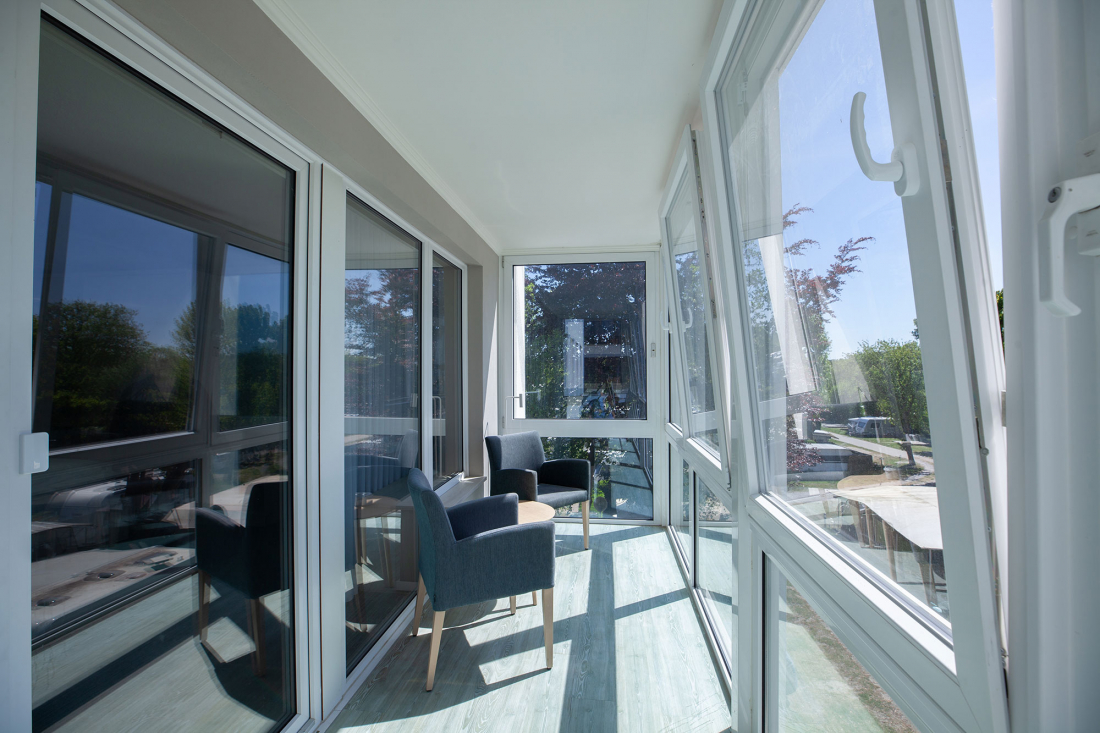 A bright, enclosed balcony with large glass windows, two modern grey chairs, a small round table and sunlight that falls on the light wooden floor. Outside you can see trees and green spaces.