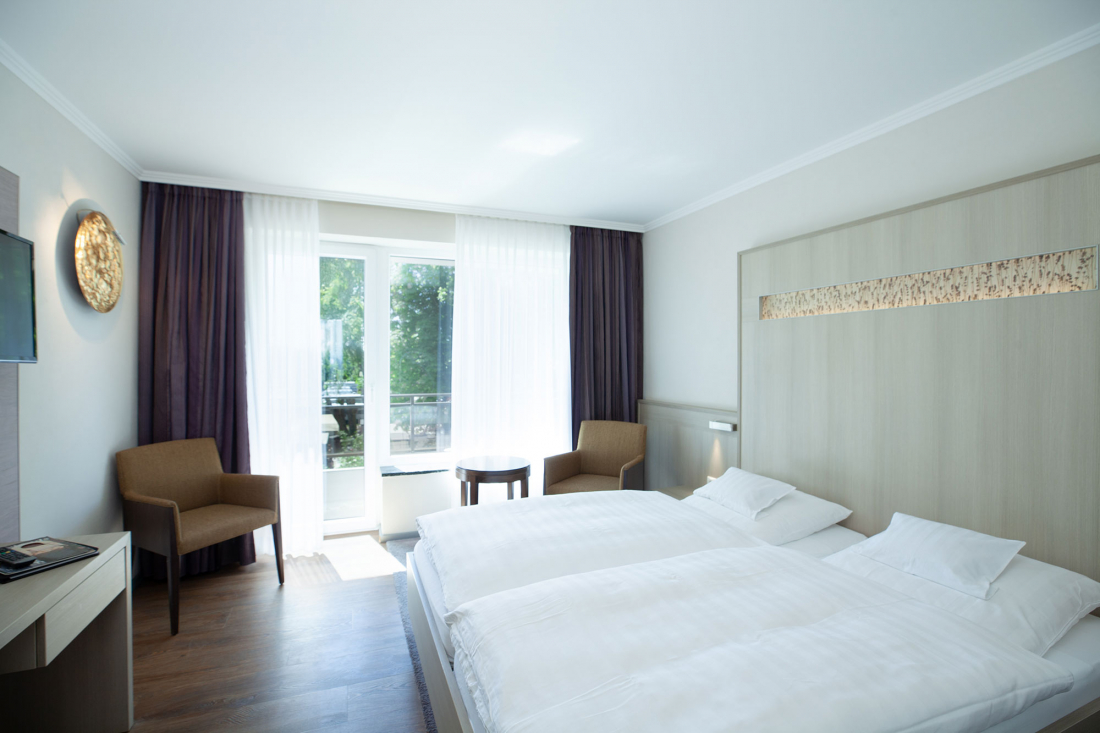 Bright hotel room with a large bed, two brown chairs, a small round table, a desk with TV and a window with transparent curtains that let in natural light. The furnishings are modern and minimalist.
