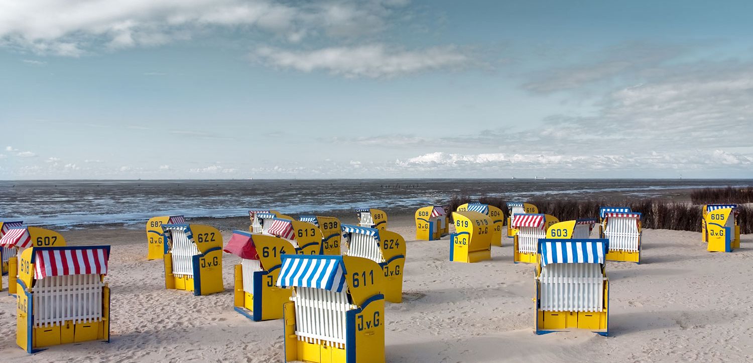 Yellow beach chairs with striped blue or red umbrellas are arranged on a sandy beach overlooking the sea under a partly cloudy sky. Each chair bears a number and the initials J.v.G.