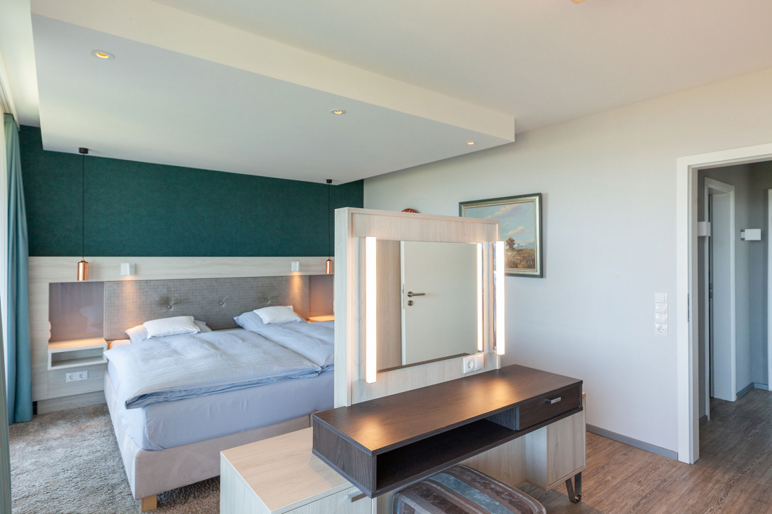 Zwei-Raum-Suite Schlafzimmer mit einem großen Bett, tealfarbener Akzentwand, Nachttischlampen, einem Waschtisch mit Spiegel und Beleuchtung, Holzboden und einer Tür, die in einen anderen Bereich führt. An der Wand hängt ein Landschaftsgemälde.