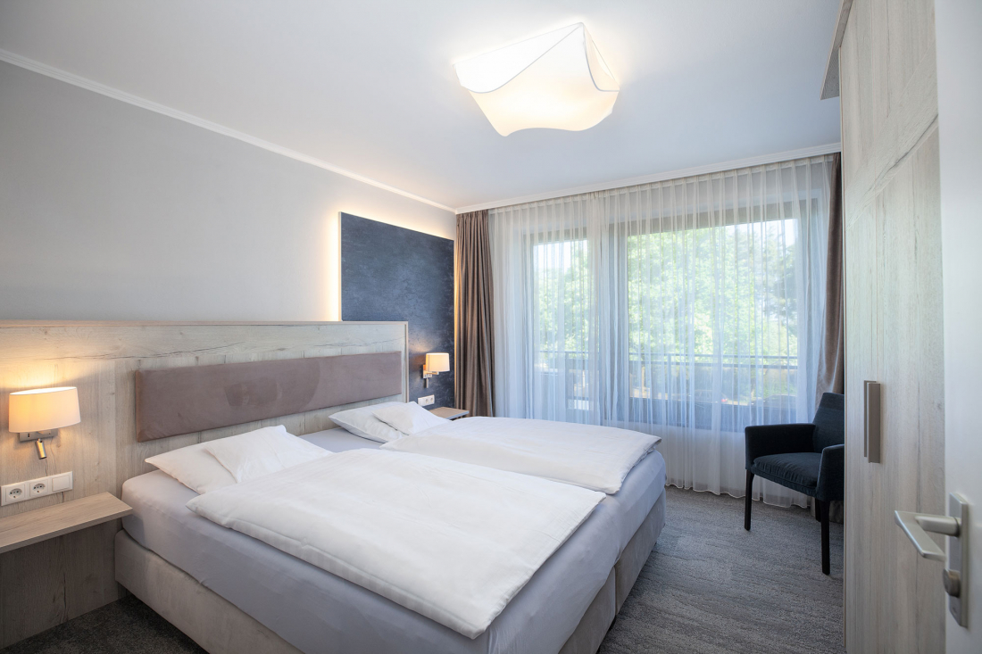 Schlafzimmer eines Dreiraum-Appartements mit Doppelbett, hellen Holzmöbeln, Nachttischlampen, einer dunklen Akzentwand, einem großen Fenster mit durchsichtigen Vorhängen und einem schwarzen Stuhl am Fenster.