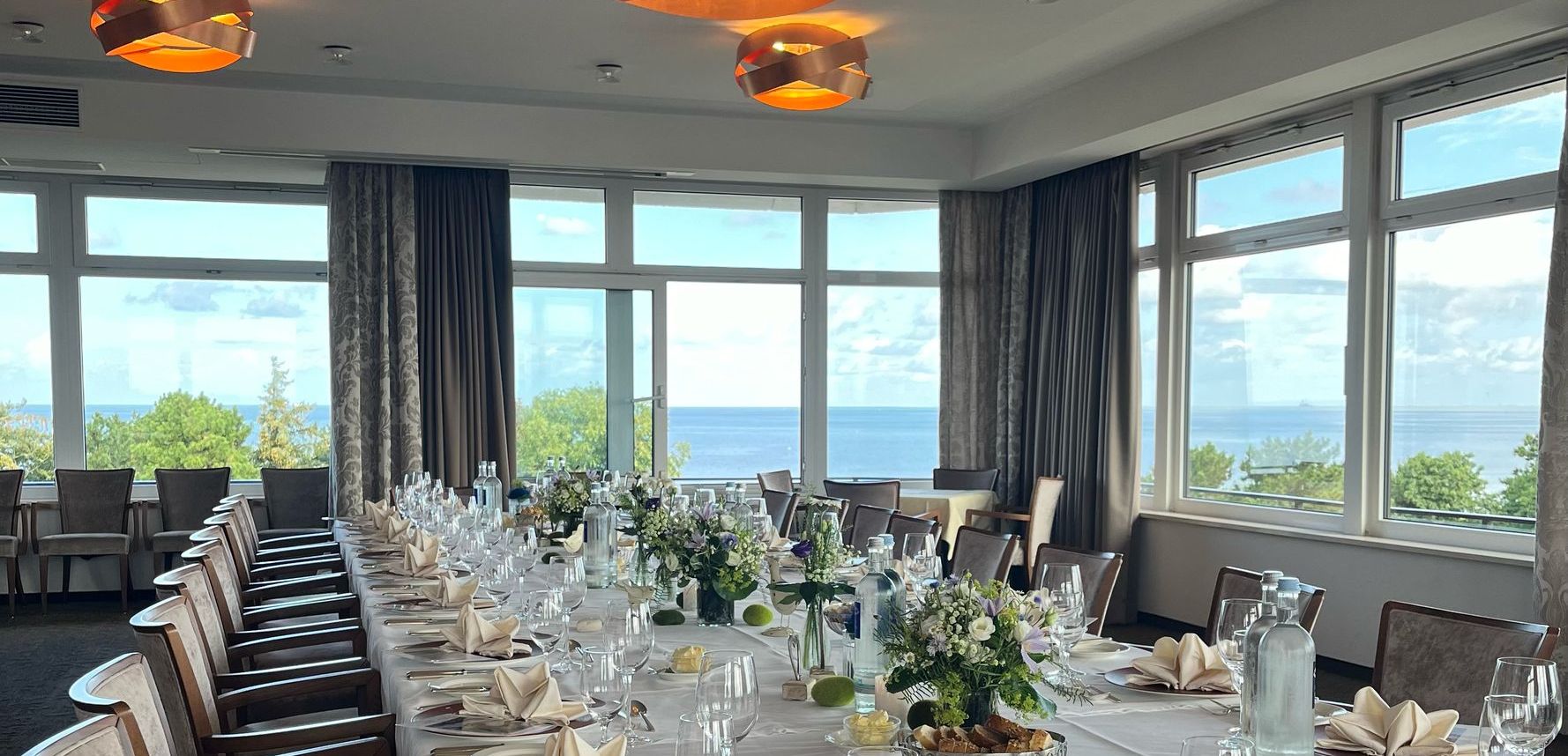 A long table set for an elegant event with white tablecloths, folded napkins, glasses, floral decorations and large windows offering a picturesque view of trees and the North Sea under a cloudless sky.