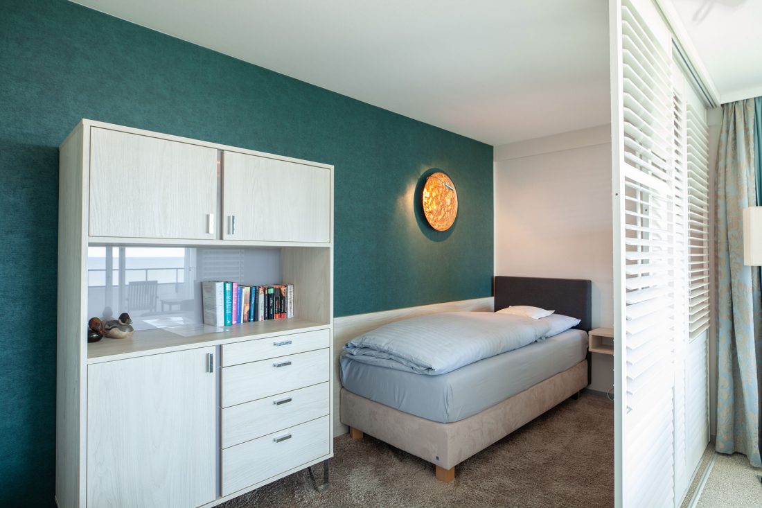 Separates Einzelbett im Wohnzimmer der Zwei-Raum-Suite, einem grauen Kopfteil, einer blauen Akzentwand, einer runden Wandleuchte und einem weißen Schrank mit Büchern und Schubladen. Ein Fenster mit weißen Jalousien lässt natürliches Licht herein.