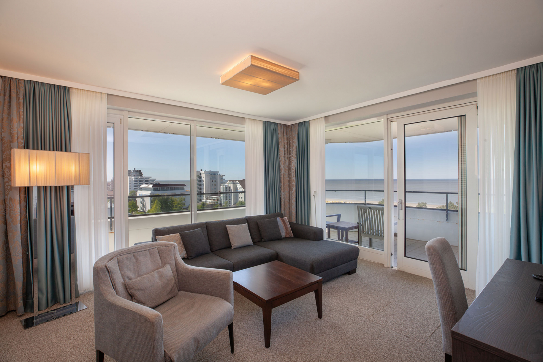 Ein modernes Wohnzimmer mit einem grauen Sektionssofa, einem Sessel, einem Couchtisch aus Holz, einer Stehlampe und großen Fenstern mit Balkonblick auf Gebäude und das Meer im Hintergrund.
