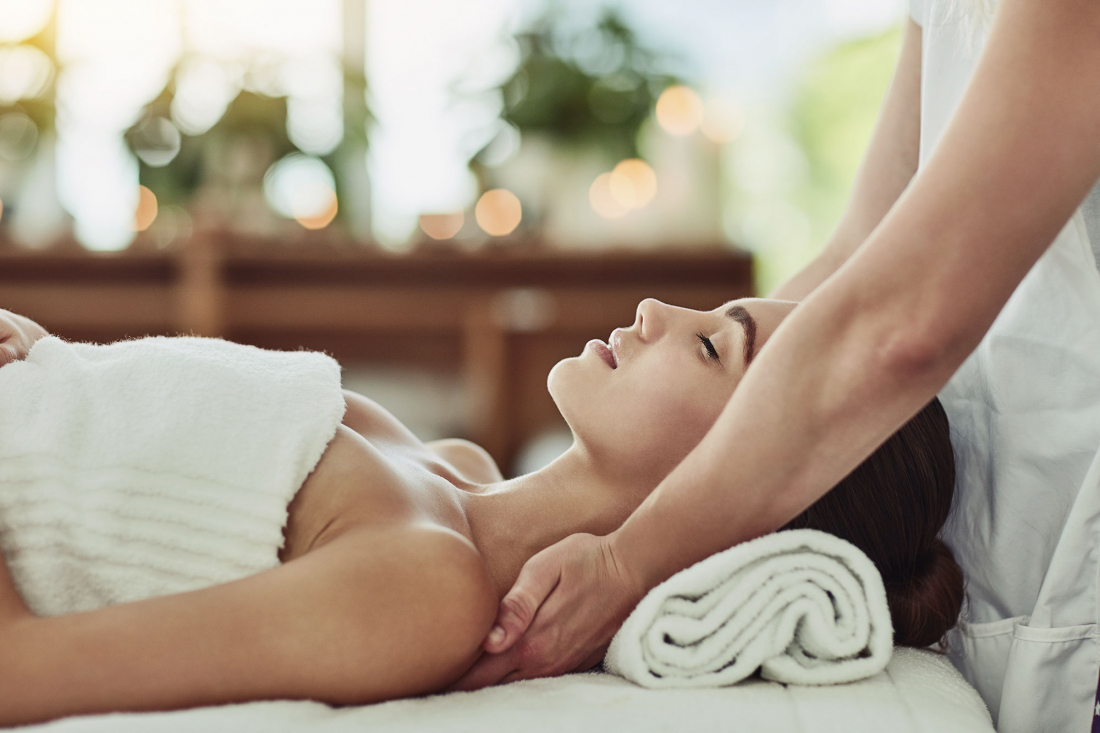 Eine Frau liegt mit geschlossenen Augen auf dem Rücken, in ein weißes Handtuch gehüllt, und erhält von einem Therapeuten eine Nackenmassage in einer friedlichen, sanft beleuchteten Spa-Umgebung mit verschwommenen Pflanzen im Hintergrund.