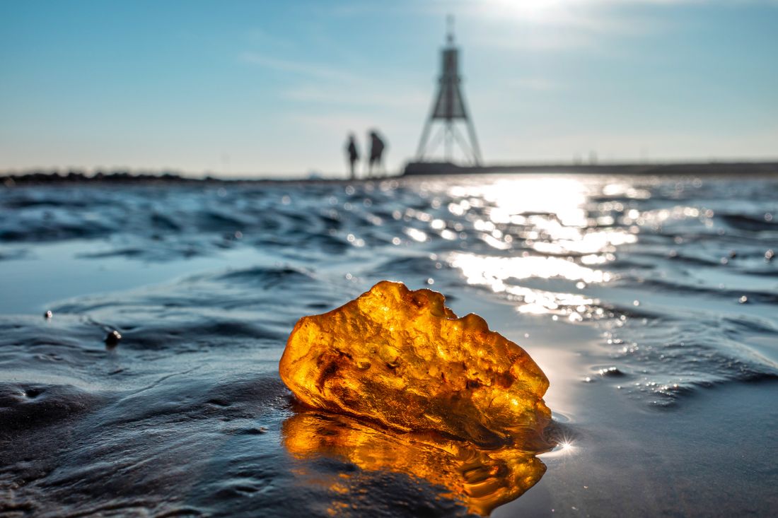 A large piece of amber lies in the wet mudflats on the coast and glistens in the sunlight. In the background, two blurred figures walk towards the Kugelbake under a bright blue sky.