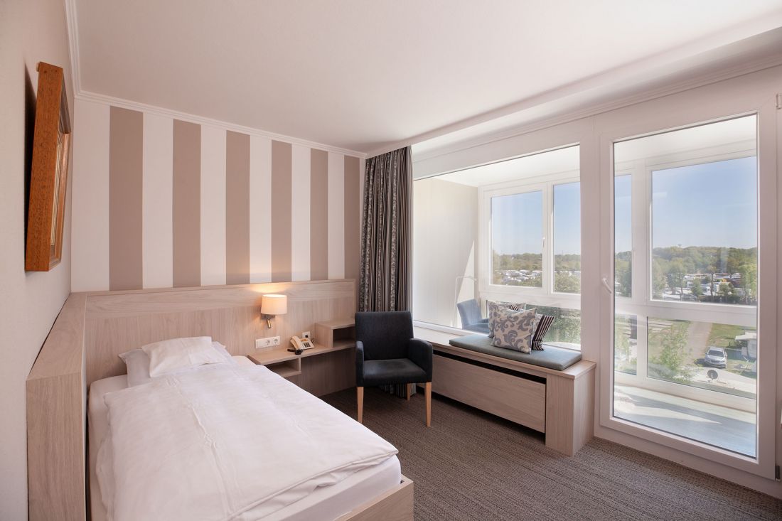 A bright, modern hotel room with a single bed, a striped accent wall, a bedside table, an armchair and a large window seat overlooking a picturesque outdoor area. The room is flooded with natural light.