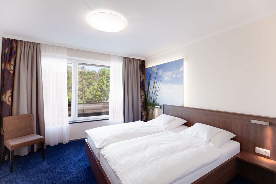 Modernes Zweiraum-Appartement mit großem Fenster, Doppelbett mit weißer Bettwäsche, blauem Teppich, braunem Stuhl und einem Kunstwerk mit Naturmotiven über dem Kopfteil. Außerhalb des Fensters sind Bäume und Grünflächen zu sehen.