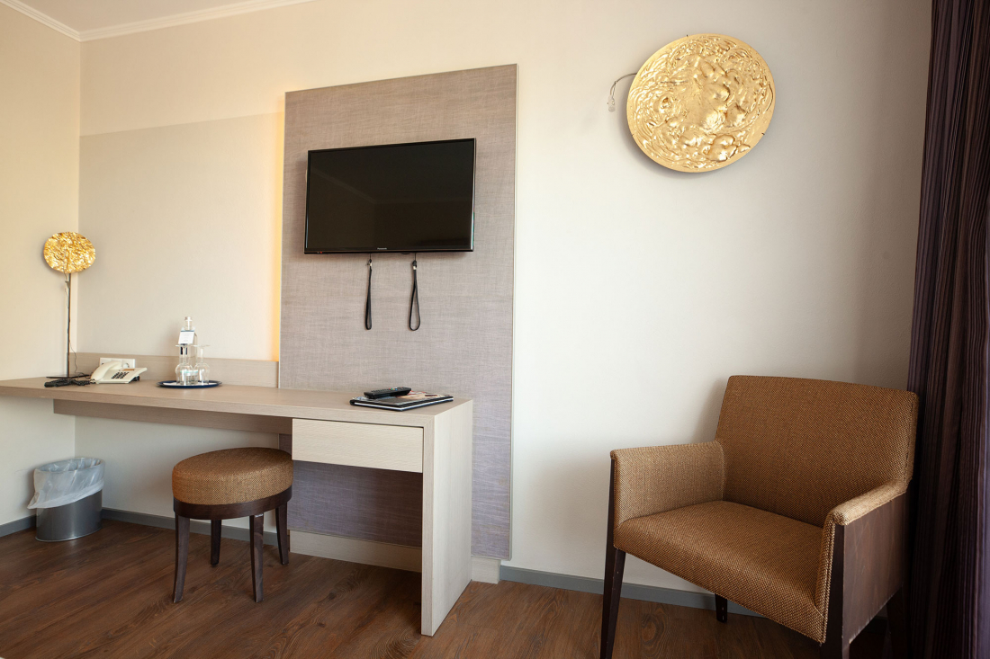 A modern small storage area in the double room with a wall-mounted TV, a desk with stool, telephone, water bottle and notepad, a brown armchair and two golden decorative wall panels.