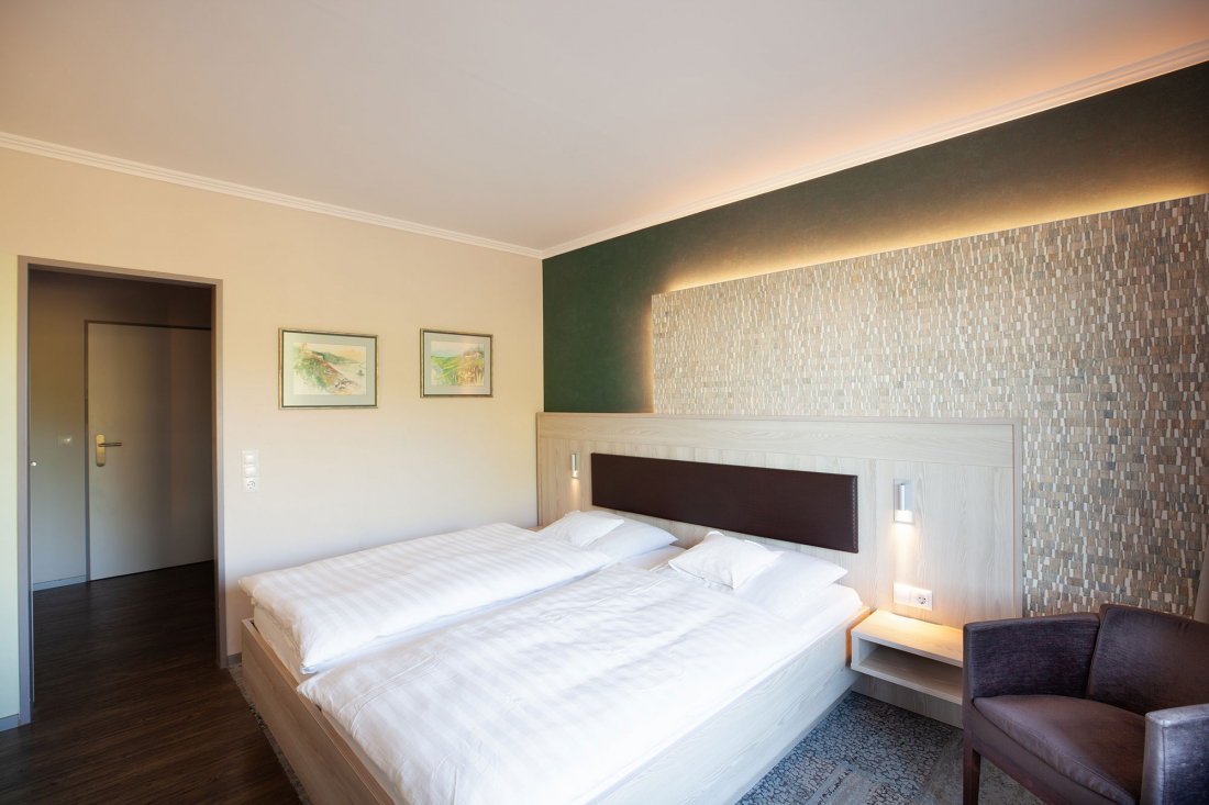 A modern hotel room with a double bed, white bedding, a patterned accent wall, two paintings, a bedside table and a purple armchair next to the bed. The room is warmly lit and has a door on the left-hand side.