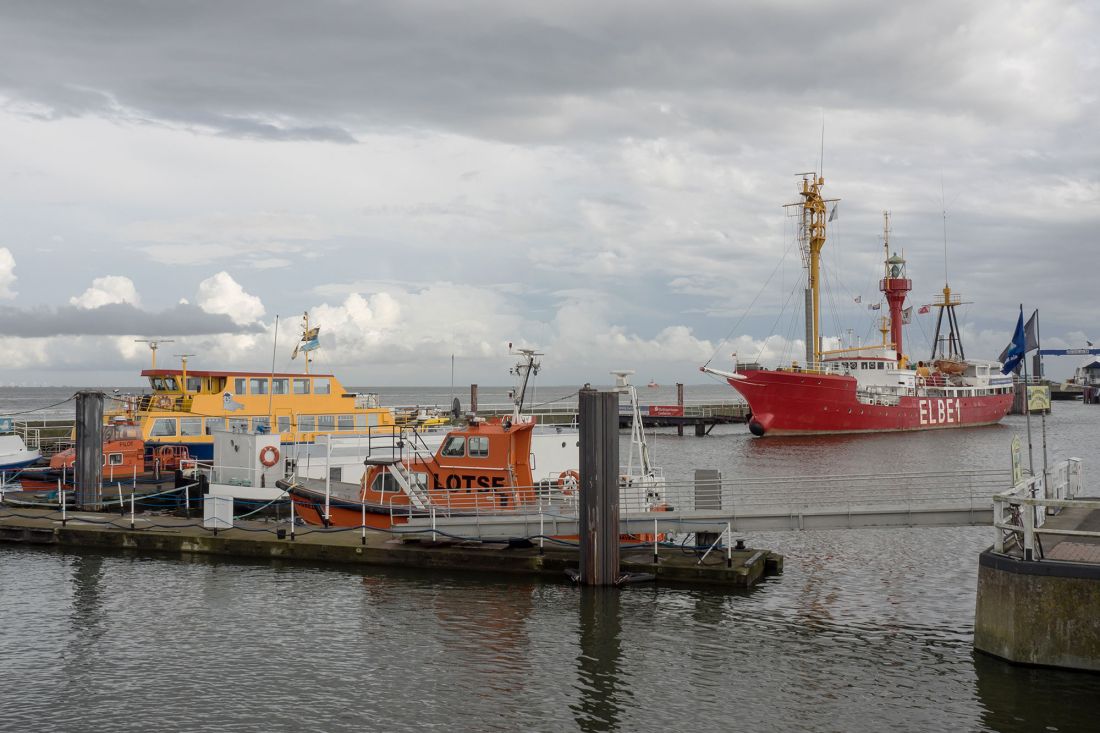 Mehrere Boote liegen im Hafen vor Anker. Im Vordergrund liegen orangefarbene und gelbe Boote an der Pier, und im Hintergrund liegt das große rotes Feuerschiff mit der Aufschrift ELBE1 vor Anker. Der Himmel ist bewölkt.