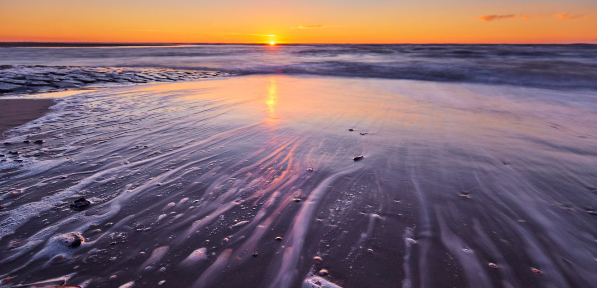 Sonnenuntergang am ruhigen Strand, an dem sanfte Wellen den nassen Sand umspülen. Der Himmel ist orange und violett und spiegelt sich im Wasser und im Sand wider, wodurch eine friedliche und beschauliche Atmosphäre entsteht.
