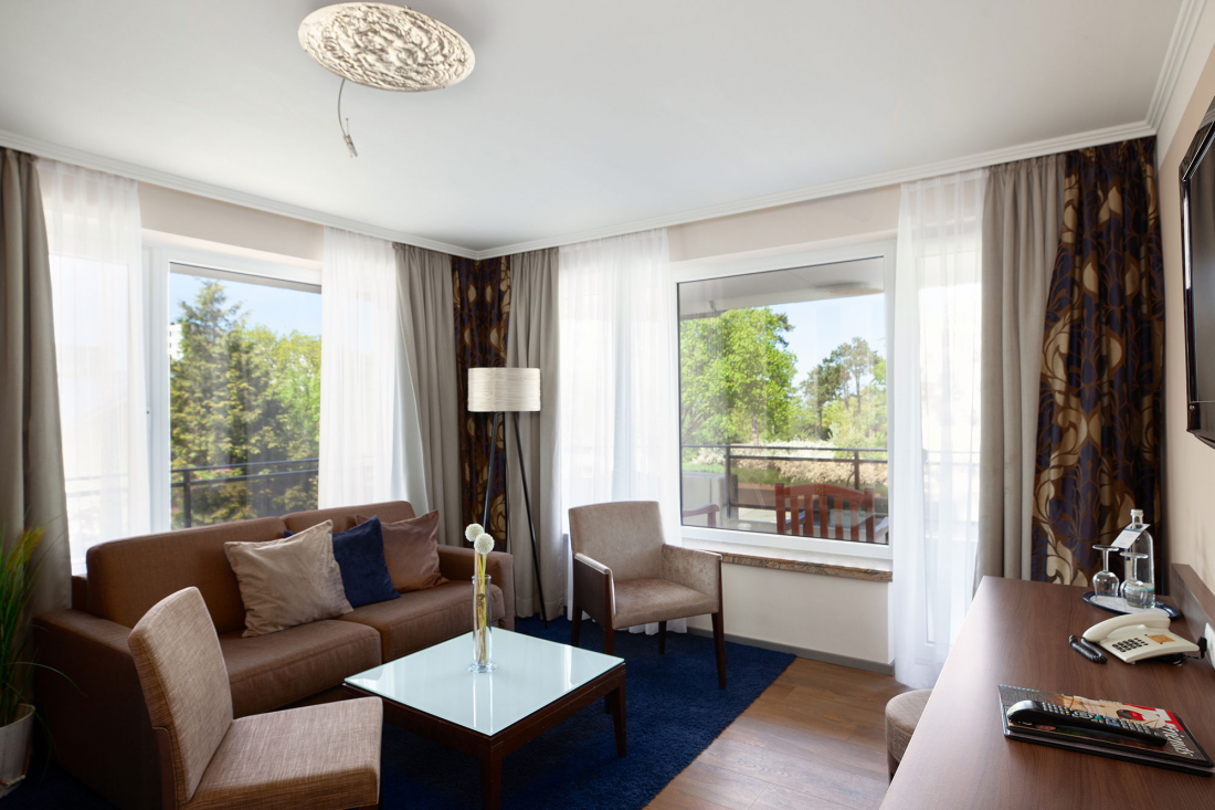 Ein modernes Wohnzimmer eines Zweiraum-Appartements mit einem braunen Sofa, einem Couchtisch aus Glas, einer Stehlampe, einem Schreibtisch und großen Fenstern mit transparenten Vorhängen, die nden Blick auf die grünen Bäume draußen freigeben.
