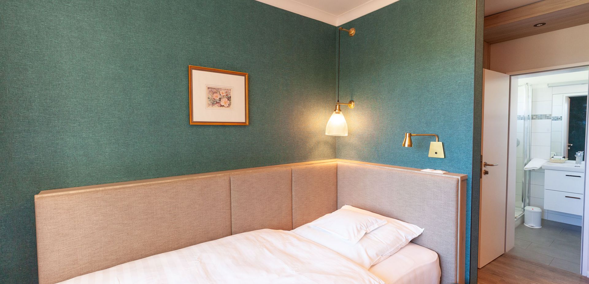 A small, modern hotel room with teal-coloured walls, a single bed with white bed linen, a wall lamp, framed artwork and an open door leading into a bright bathroom.