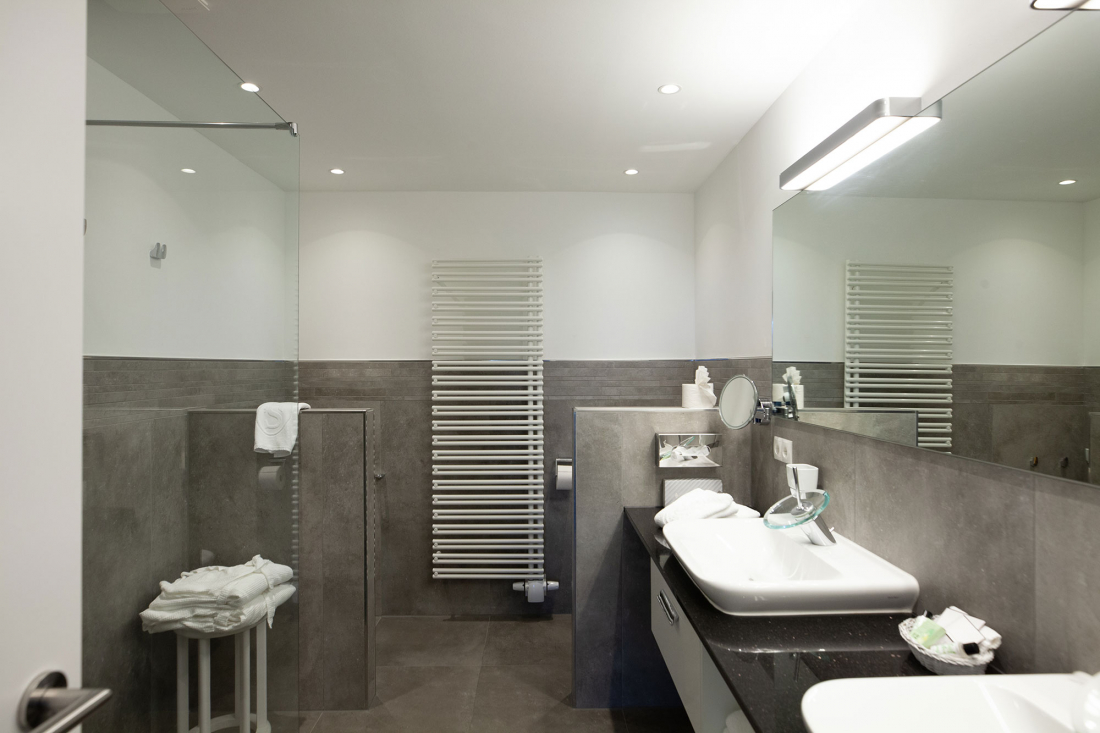 Modernes Badezimmer mit grauen Fliesen, einem großen Spiegel über einem weißen Waschbecken auf einer schwarzen Arbeitsplatte, einer gläsernen Duschabtrennung, einem weißen Handtuchhalter und ordentlich gefalteten Handtüchern auf einem kleinen Hocker.