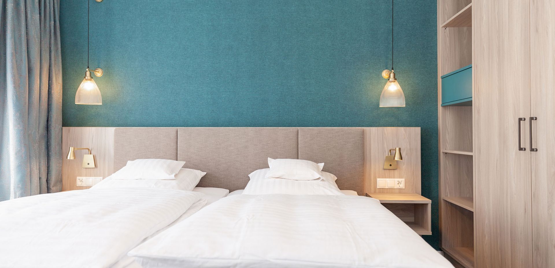 Two single beds with white bed linen stand side by side against a blue-green accent wall, each with a pillow and a reading lamp attached to the wall. Pendant lights hang above the wooden bedside tables on either side of the beds.