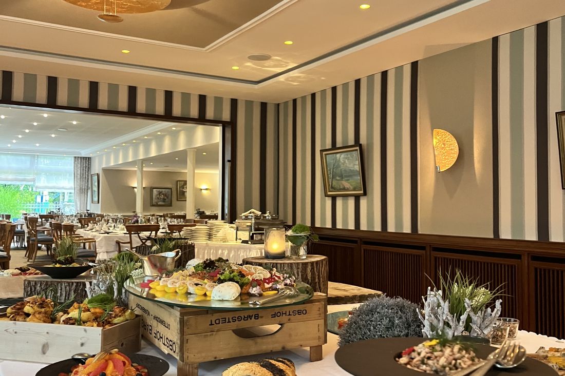 A buffet table with various dishes, bread, and salads on wooden stands in a stylish dining room with striped walls, framed art, and a set dining area in the background.