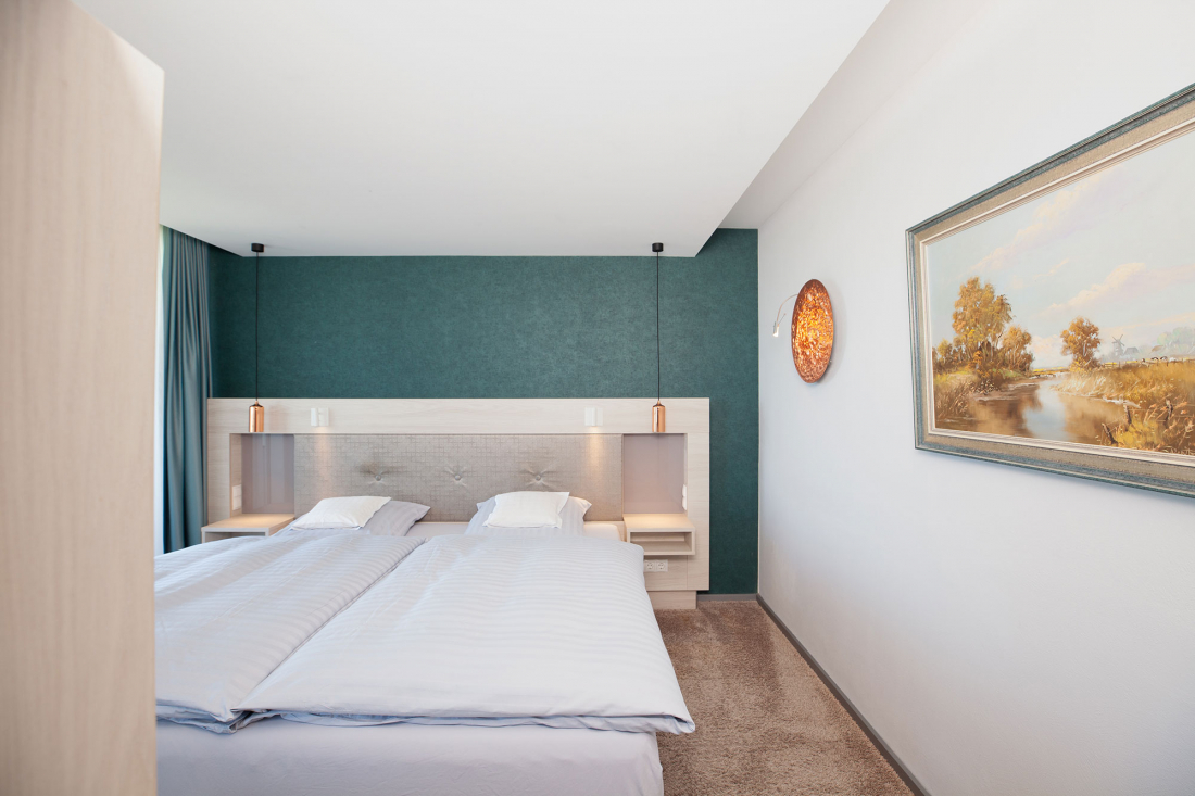Ein modernes, minimalistisches Schlafzimmer mit Doppelbett, tealfarbener Akzentwand, weißer Bettwäsche und einem Landschaftsgemälde an einer weißen Wand. Über den Betten hängen kupferfarbene Beleuchtungskörper.
