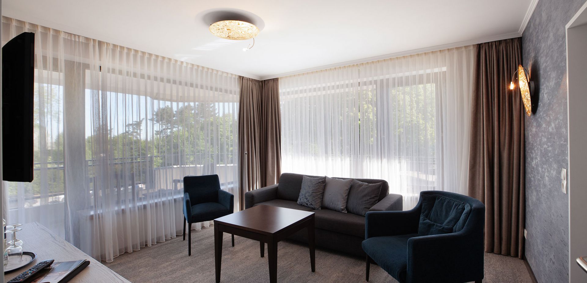 Ein helles Wohnzimmer eines Dreiraum-Appartement mit durchsichtigen Vorhängen, grauem Sofa mit Kissen, zwei dunkelblauen Sesseln und einem kleinen Couchtisch aus Holz. Große Fenster lassen natürliches Licht herein.