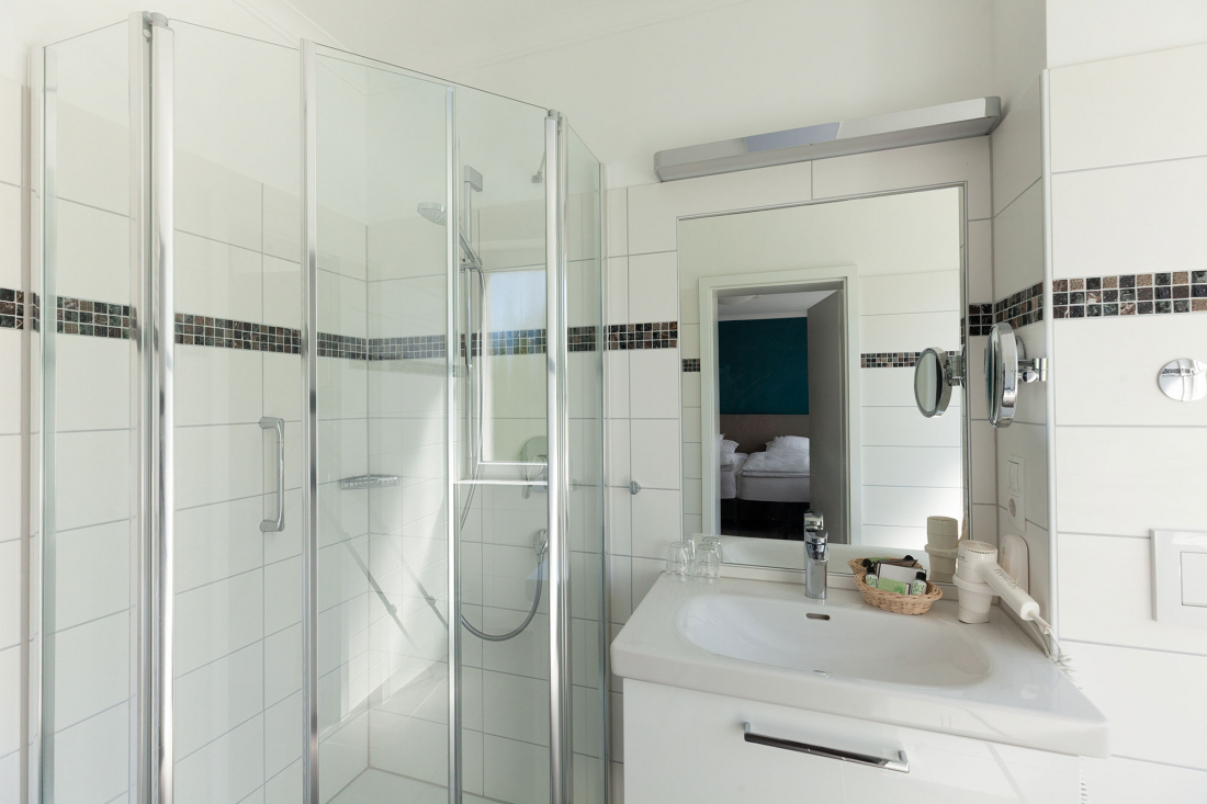 Modern bathroom with white tiled walls, a glazed corner shower, a white washbasin with a mirror above and a hairdryer on the counter. An open door provides a view of a bedroom in the background.