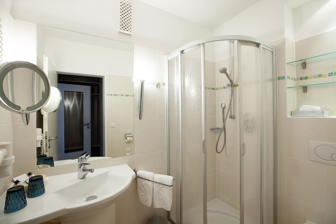 A modern, clean bathroom with a glass shower cubicle, a washbasin with mirror, wall shelves, towels and blue toilet cups on the counter. The walls are tiled in a light beige colour.