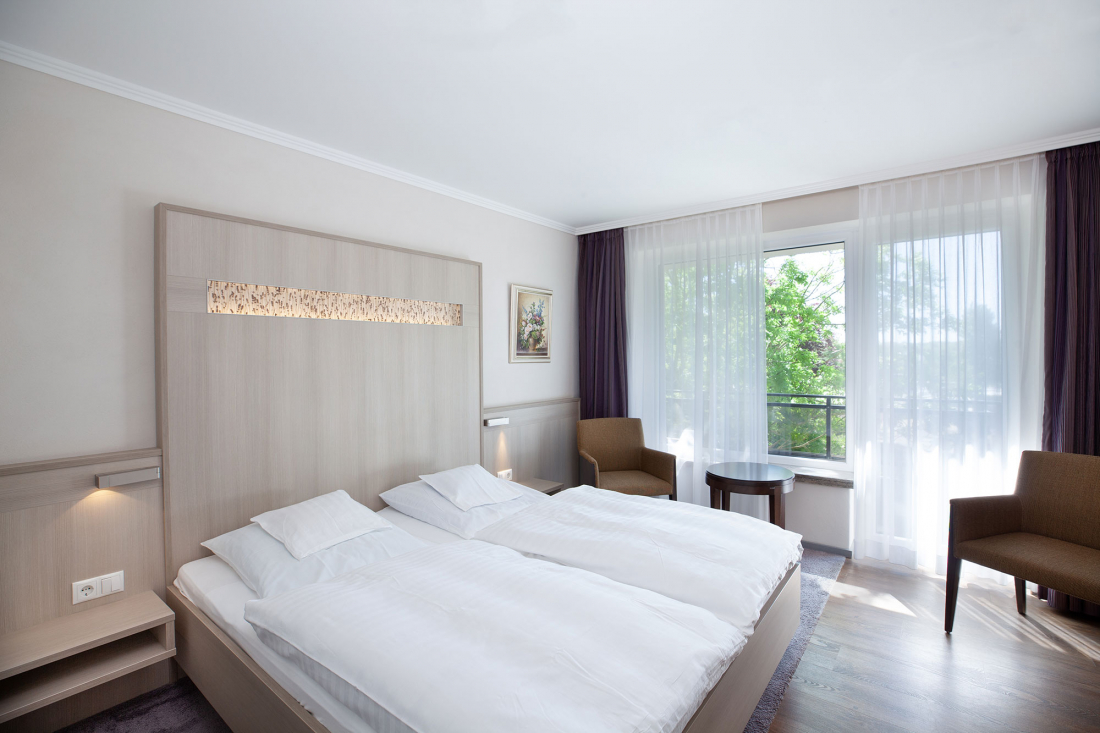 A bright, modern hotel room with two single beds pushed together, white bedding, a wooden headboard, two brown chairs at a round table, a large window with sheer curtains and a painting on the wall.