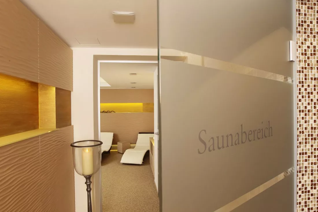 A frosted glass door labeled "Sauna Area" leads into a modern sauna relaxation area with lounge chairs, warm lighting, and textured walls. A tall candlestick stands next to the entrance.