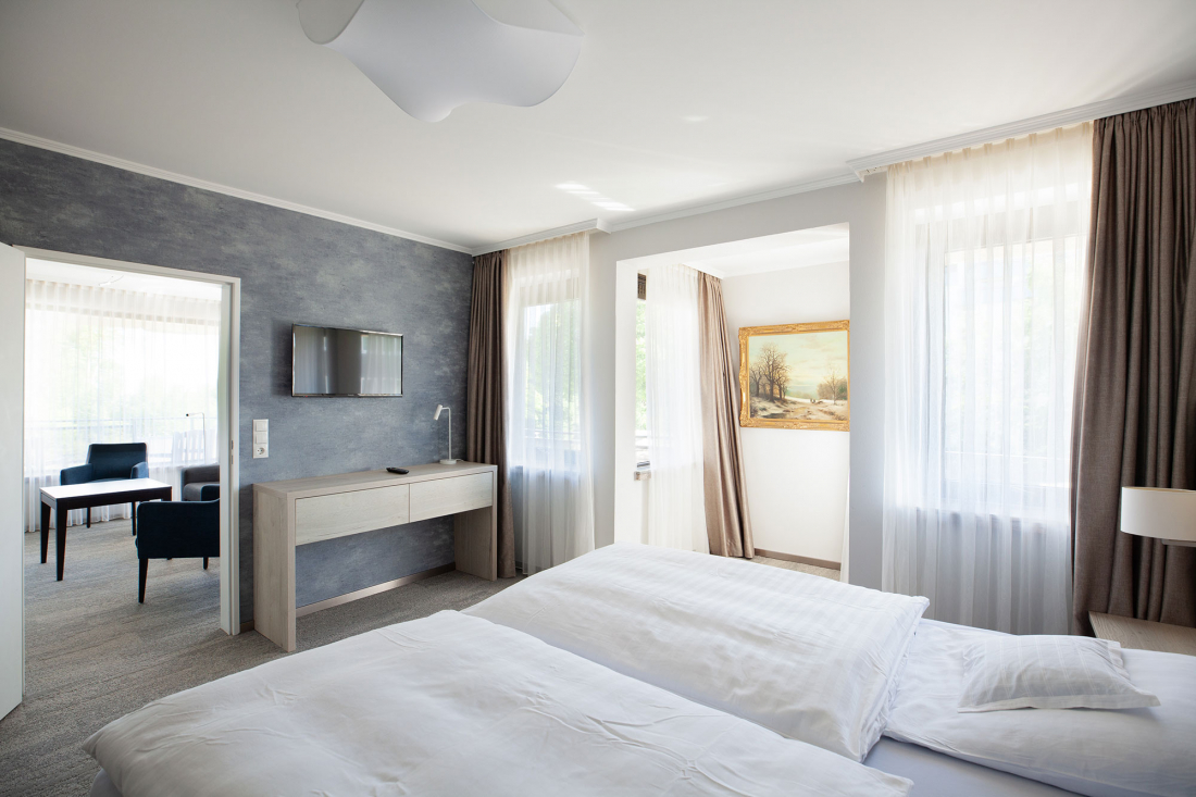 Ein helles, modernes Appartementzimme mit einem großen Bett, einem Schreibtisch mit Stuhl, einem an der Wand montierten Fernseher und großen Fenstern mit transparenten Vorhängen. Eine angrenzende Sitzecke und ein gerahmtes Gemälde sind ebenfalls zu sehen.