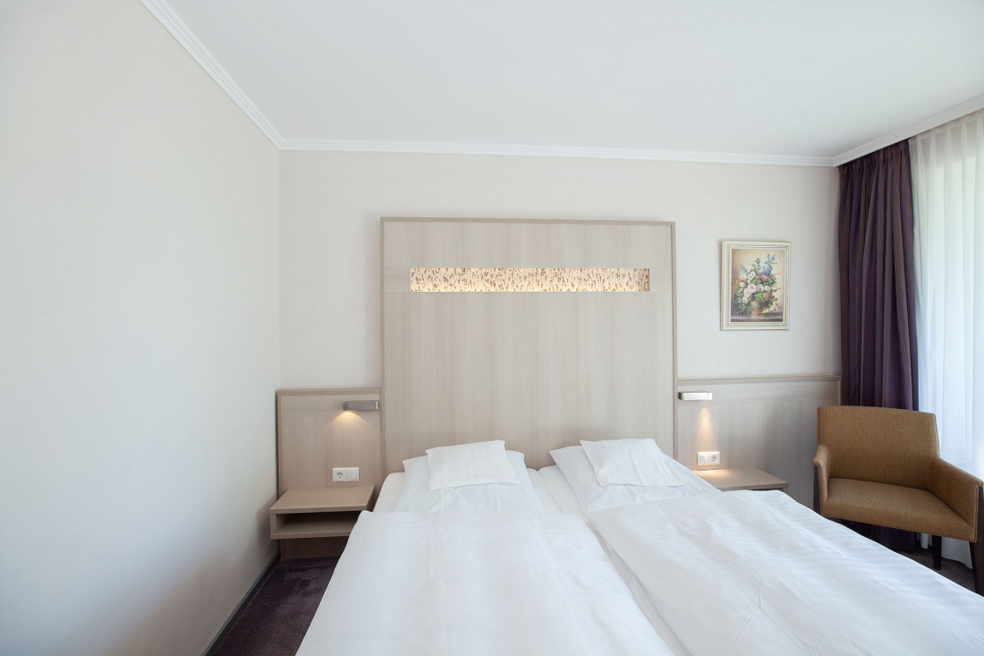 A double room with a double bed, white bedding, a light wooden headboard with built-in lamps, a bedside table on each side, an armchair, a framed picture and a large window with sheer curtains.
