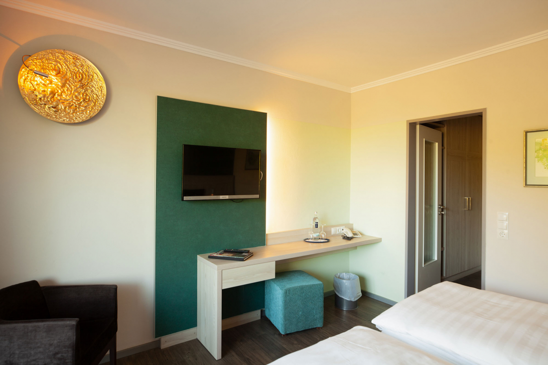 A modern double room with a wall-mounted television above a light wood desk, a teal ottoman, a chair, a bright round wall lamp, two beds, and an open door leading to a hallway.