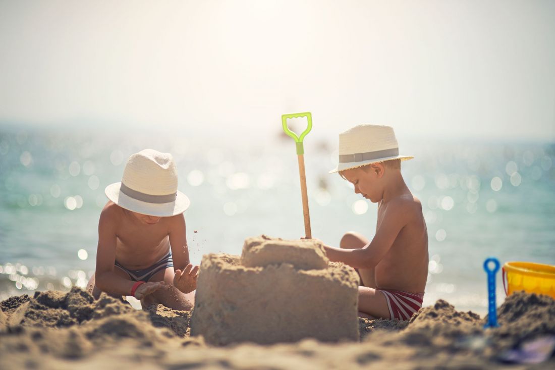Two little boys wearing straw hats are building a sandcastle on a sunny beach. One boy is pounding sand, while the other is concentrating on the castle, from which a green shovel protrudes. The sea can be seen in the background.