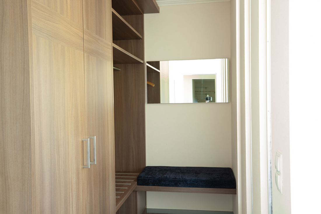 Ein kleiner abgetrenter Kleiderschrank mit Holzregalen, einer Kleiderstange, einer eingebauten Bank mit einem dunklen Kissen und einem rechteckigen Spiegel an der Wand, der einen Teil des Raumes reflektiert.
