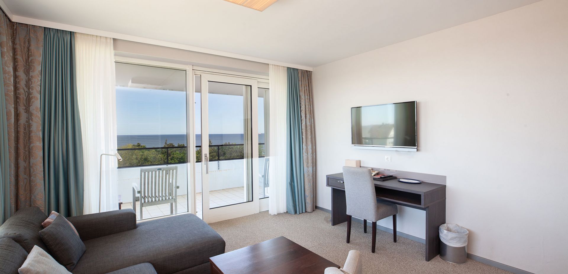 Helles Wohnzimmer der Zwei-Raum-Suite mit einem Sofa, einem Couchtisch, einem Schreibtisch und einem an der Wand montierten Fernseher. Große Glastüren führen auf einen Balkon mit Stühlen und bieten einen Blick auf das Meer und das Grün draußen.