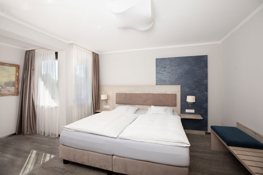 Ein modernes Hotelzimmer des Drei-Raum-Apartments mit einem großen Doppelbett, weißem Bettzeug, zwei Nachttischen mit Lampen, einer blauen Akzentwand, großen Fenstern mit Vorhängen und einer Bank an einer Wand.