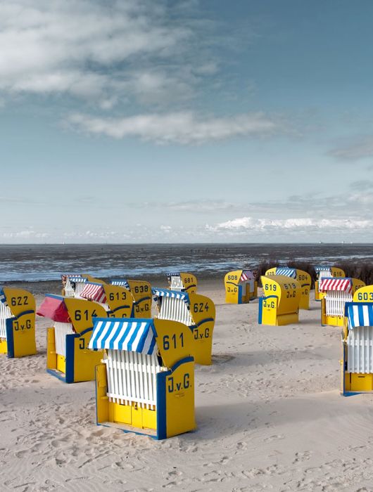 Gelbe Strandkörbe mit gestreiften blauen oder roten Schirmen sind an einem Sandstrand mit Blick auf das Meer unter einem teilweise bewölkten Himmel angeordnet. Jeder Stuhl trägt eine Nummer und die Initialen J.v.G.