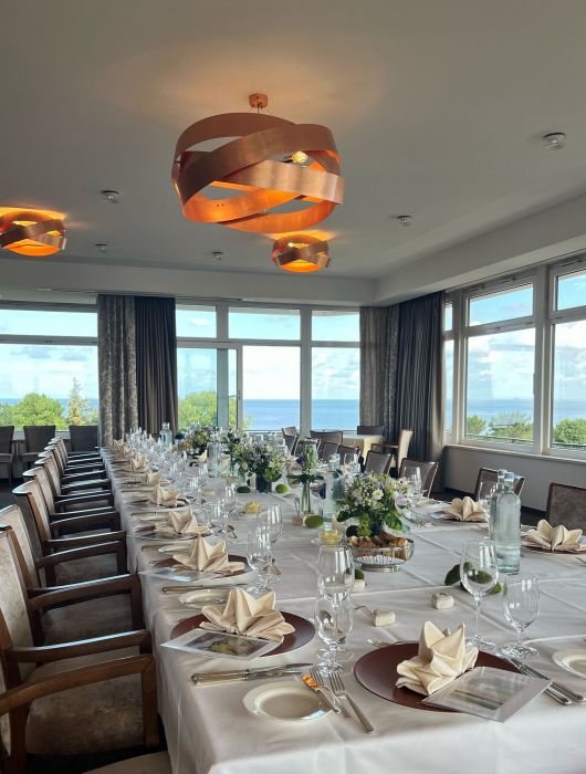 Ein langer, für eine elegante Veranstaltung gedeckter Tisch mit weißen Tischtüchern, gefalteten Servietten, Gläsern, Blumenschmuck und großen Fenstern, die einen malerischen Blick auf Bäume und die Nordsee unter einem wolkenfreien Himmel bieten.