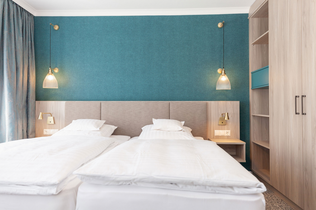 Two single beds with white bed linen stand side by side against a blue-green accent wall, each with a pillow and a reading lamp attached to the wall. Pendant lights hang above the wooden bedside tables on either side of the beds.