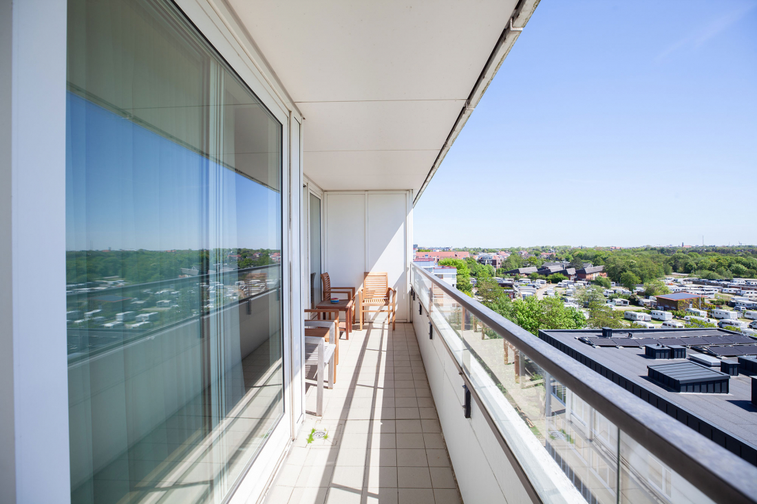 Ein langer, schmaler Balkon mit Glasgeländer und Holzstühlen überblickt den Campingplatz mit Bäumen und Gebäuden unter einem klaren blauen Himmel.