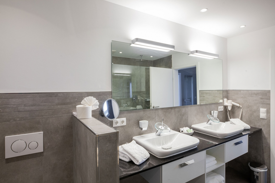 Modernes Badezimmer mit zwei quadratischen Waschbecken, einem großen Wandspiegel und grauen Fliesenakzenten. Handtücher, Toilettenpapier und Toilettenartikel sind ordentlich auf der Arbeitsplatte unter der hellen Deckenbeleuchtung angeordnet.