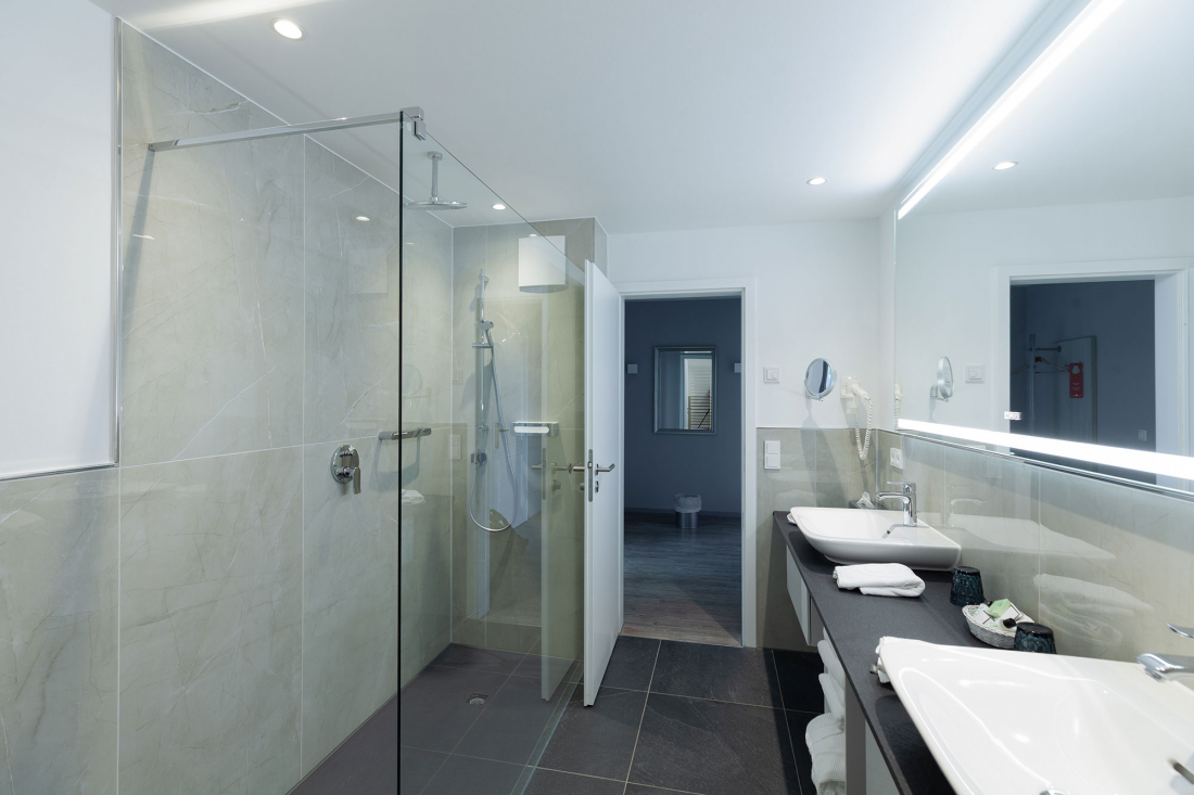 Modernes Badezimmer mit Glasduschkabine, Doppelwaschbecken auf einer schwarzen Arbeitsplatte, großem Spiegel mit integrierter Beleuchtung und hellen Wandfliesen. Handtücher und Toilettenartikel sind ordentlich auf dem Tresen angeordnet.