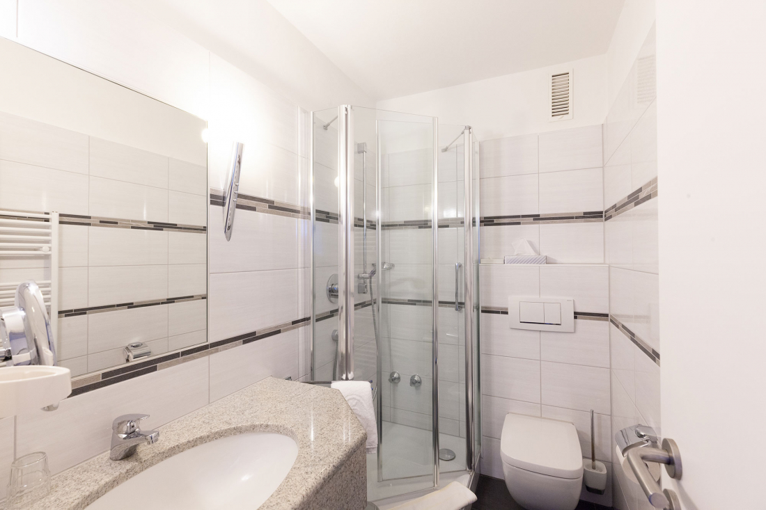 Modern white bathroom with tiled walls, a corner shower with glass screen, a wall-mounted toilet, a washbasin with granite worktop and a large mirror above the washbasin.