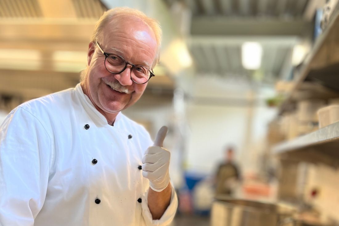 Jörg Hansen, wearing glasses and a mustache, gives a thumbs-up in a busy kitchen. He's wearing a white chef's jacket and disposable gloves. The background is blurred, showing kitchen appliances and another person.