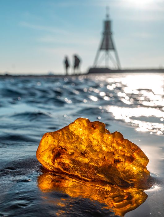 Ein großes Stück Bernstein liegt im nassen Watt an der Küste und glitzert im Sonnenlicht. Im Hintergrund gehen zwei unscharfe Gestalten unter strahlend blauem Himmel auf die Kugelbake zu.