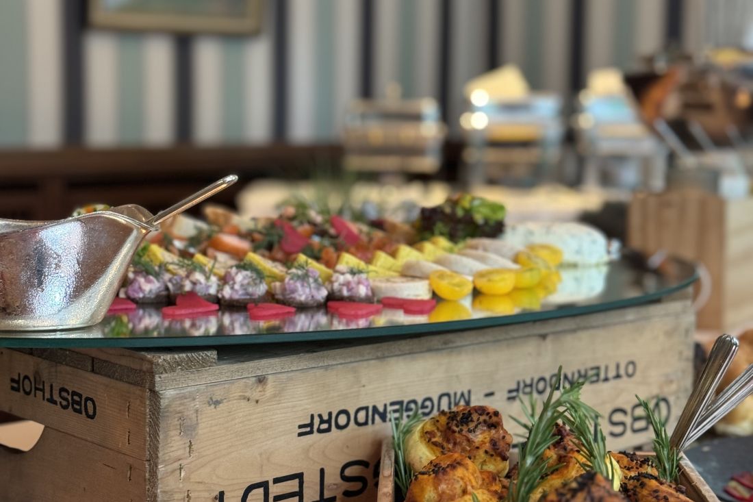 Close-up of a buffet table with a selection of colorful appetizers and cheeses on a glass surface displayed on a wooden box, with serving utensils and a blurred background.