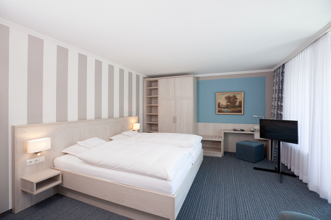 A modern hotel room with a double bed, striped wallpaper, bedside lamps, a wardrobe, a desk with TV and a large window with sheer curtains that let in natural light.